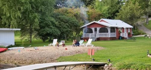 A cozy red cottage by a sandy beach, with people sitting on chairs and smoke rising from a nearby fire.