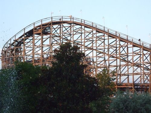 A wooden roller coaster rises against a clear sky, surrounded by trees and a water feature in the foreground.