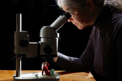 A woman examines a small figure under a microscope, illuminated against a dark background.