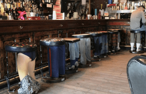 A row of unique bar stools with painted legs, resembling people in casual attire, in a rustic bar setting.