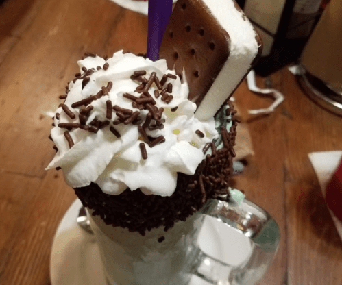 A tall milkshake topped with whipped cream, chocolate sprinkles, and an ice cream sandwich.