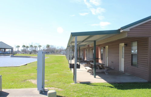 A row of buildings with a covered porch, overlooking a grassy area and a body of water under a clear blue sky.