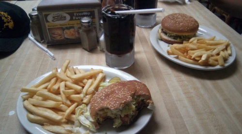 Two plates with burgers, fries, and drinks on a wooden table. One burger is partially eaten.