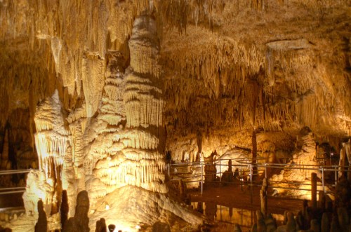 A dimly lit cave interior featuring impressive stalactites and stalagmites, with a walkway for visitors.