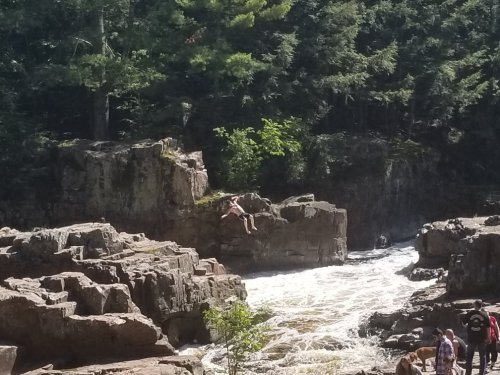 A person sits on a rock by a rushing river, surrounded by trees and rocky terrain.