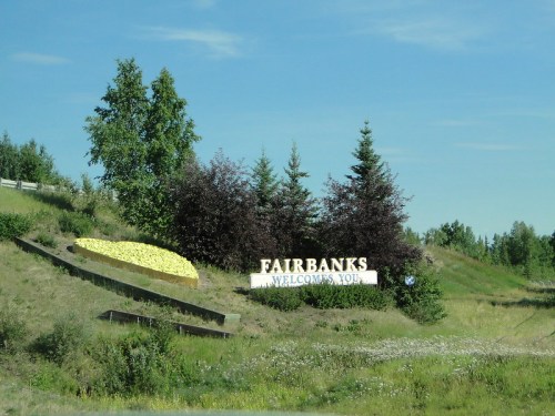 Sign welcoming visitors to Fairbanks, Alaska, surrounded by greenery and trees on a sunny day.