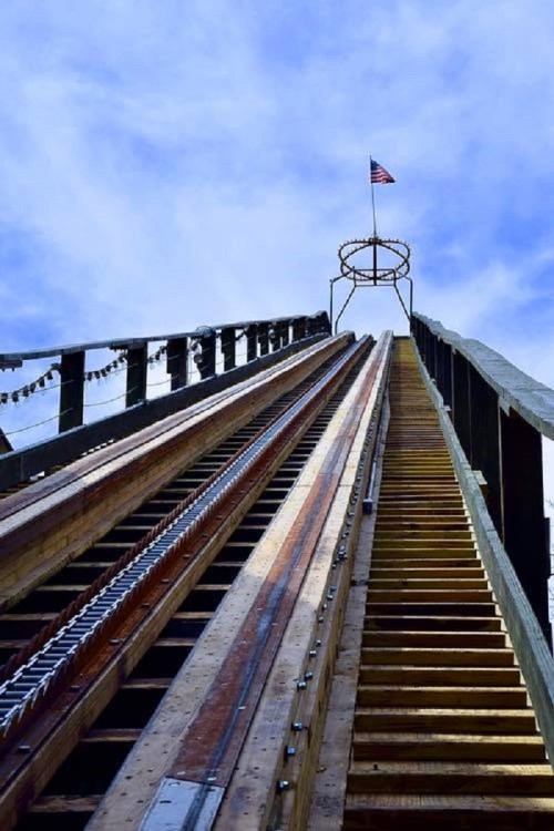 A steep wooden roller coaster track leading up to a circular loop, with an American flag at the top against a blue sky.