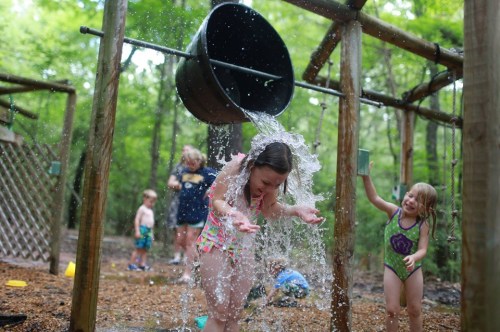 A girl in a swimsuit gets drenched with water from a bucket at a playground, while other children play nearby.