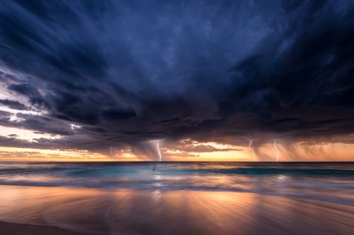 A dramatic seascape at sunset with dark clouds, lightning, and gentle waves reflecting the colorful sky.