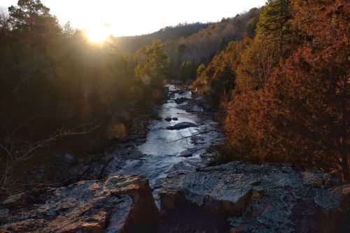 Sunset over a serene river flowing through a forested landscape, with rocky banks and trees lining the water.