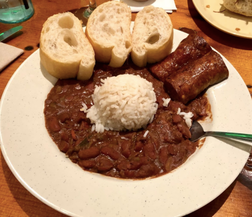 A plate of rice topped with red beans, served with slices of bread and a sausage link on the side.