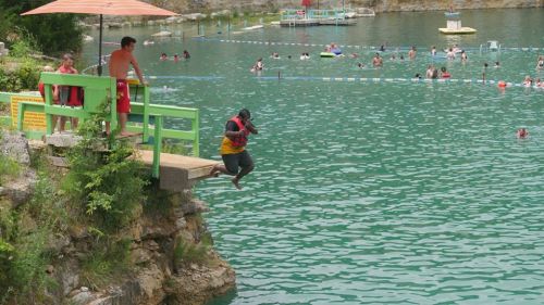 A person in a life jacket jumps off a diving platform into a turquoise swimming area, with people swimming nearby.