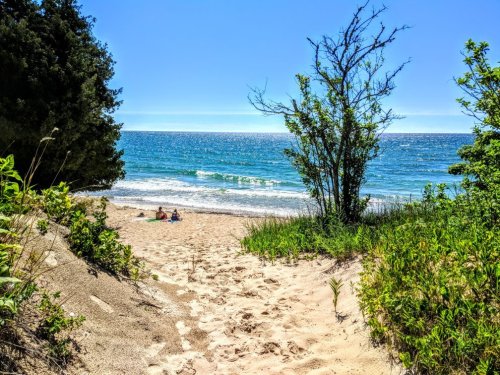 A sandy path leads to a tranquil beach with gentle waves and clear blue skies, surrounded by greenery.