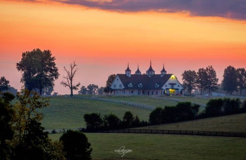 A picturesque farm at sunset, featuring a large barn with spires, surrounded by rolling hills and trees.