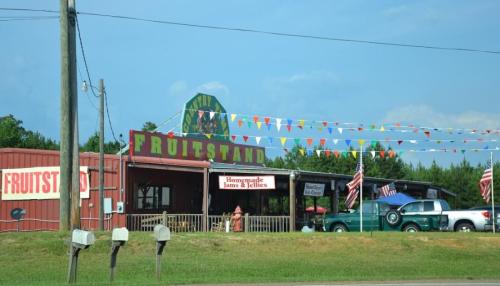 A colorful fruit stand with flags and American flags, featuring a sign that reads "Fruitstand" and vintage cars nearby.