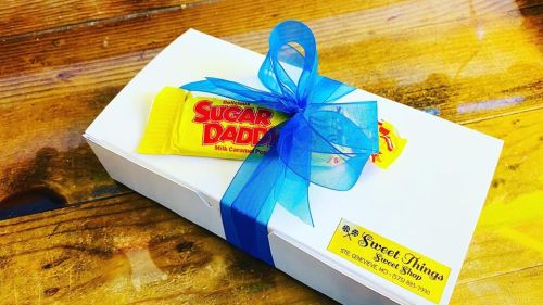 A white gift box tied with a blue ribbon, featuring a Sugar Daddy candy bar on top.