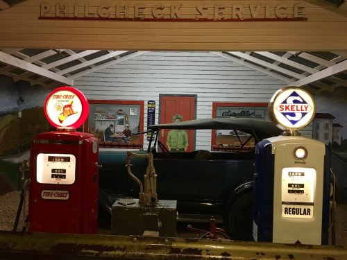 Vintage gas station with two old-fashioned pumps, a classic car, and a mural in the background.