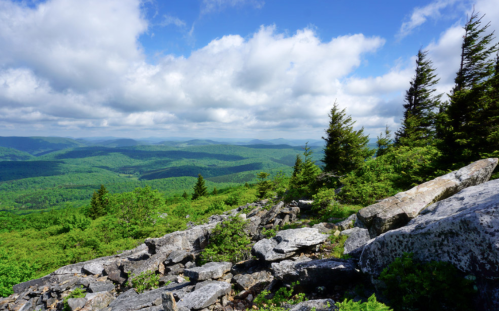A scenic view from a rocky overlook, showcasing lush green mountains and a blue sky with fluffy clouds.
