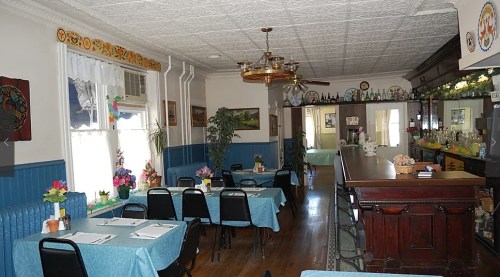 Cozy restaurant interior with blue tables, black chairs, plants, and a wooden bar, featuring decorative wall art.