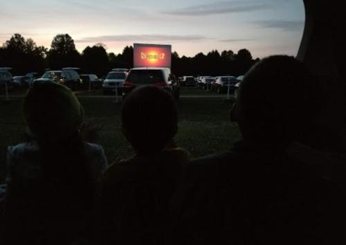 Three children watching a movie on a large screen at a drive-in theater, with cars parked in the background at dusk.