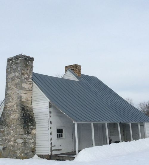 A rustic house with a gray metal roof and stone chimney, surrounded by snow under a cloudy sky.