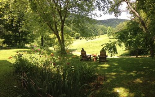 A serene garden scene with two wooden chairs, colorful flowers, and a lush green landscape under a clear sky.