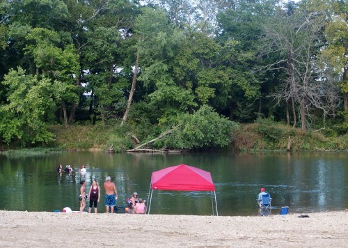 People enjoying a beach by a river, with a red canopy and lush greenery in the background.