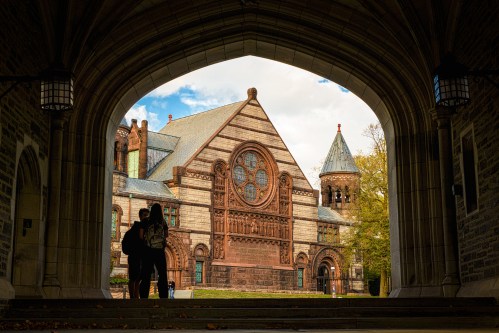 Silhouetted figures stand under an archway, framed by a historic building with intricate architecture and a cloudy sky.