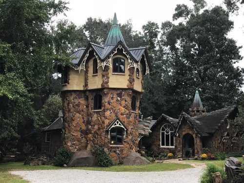 Whimsical stone cottage with a turret, green roof, and decorative windows, surrounded by trees and a gravel path.