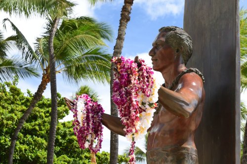A bronze statue of a man holding flower leis, surrounded by palm trees and a blue sky.
