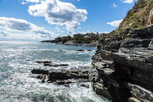 A rocky coastline under a blue sky with fluffy clouds, waves crashing against the shore.