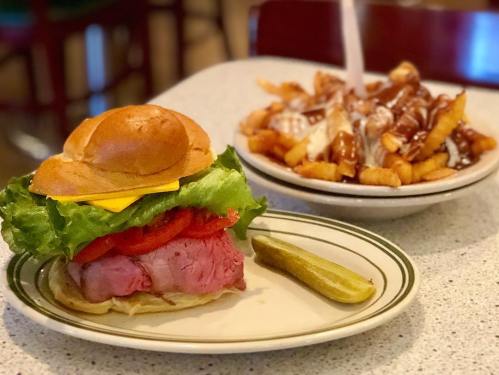 A plate with a roast beef sandwich topped with lettuce, tomato, and cheese, alongside a bowl of loaded fries.