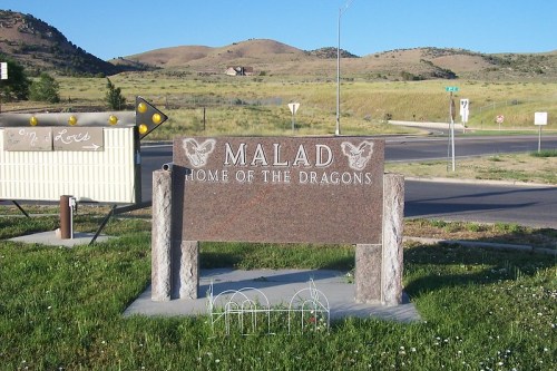 Sign for Malad, Idaho, stating "Home of the Dragons," with a scenic background of hills and grass.