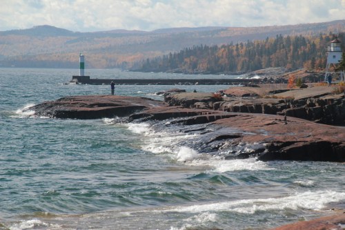 A rocky shoreline with gentle waves, a lighthouse in the distance, and autumn foliage on the hills.