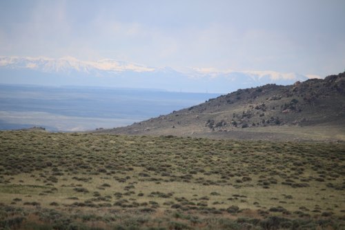 A vast landscape featuring rolling hills, sparse vegetation, and distant snow-capped mountains under a cloudy sky.