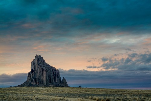 A dramatic rock formation rises against a colorful sky at sunset, surrounded by open plains.