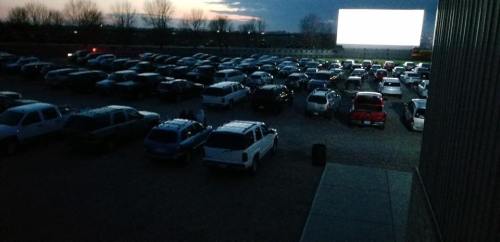 A drive-in movie theater at dusk, with numerous cars parked and a large screen displaying a film.