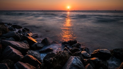 A serene sunset over the ocean, with golden light reflecting on the water and rocky shoreline in the foreground.