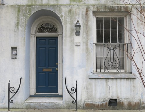 A weathered building facade featuring a blue door, a window with bars, and a light fixture.