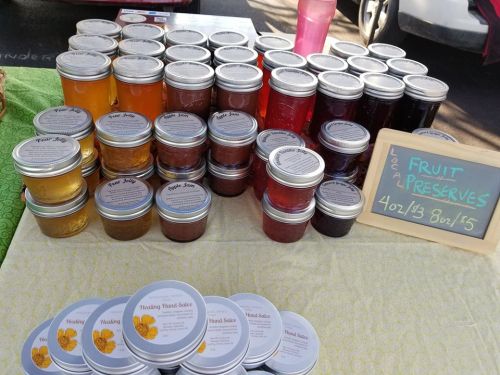 A display of various fruit preserves in jars, with a sign reading "Local Fruit Preserves" and healing hand salve products.