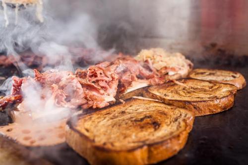 Sizzling grilled sandwiches with layers of meat and melted cheese, surrounded by steam on a hot grill.