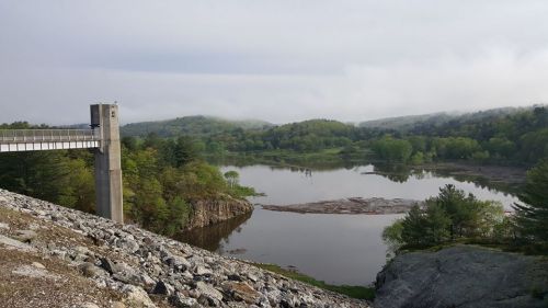 A serene landscape featuring a dam, calm water, and lush green hills under a cloudy sky.