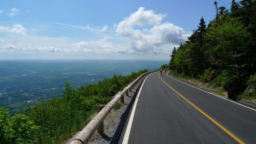 A winding road through lush greenery, with a scenic view of valleys and mountains under a blue sky with clouds.