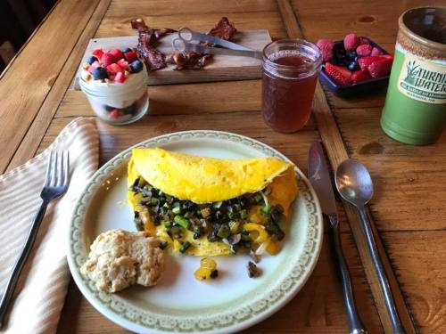 A plate with a cheese and vegetable omelet, biscuit, and a glass of iced tea, alongside fruit and bacon on a wooden table.
