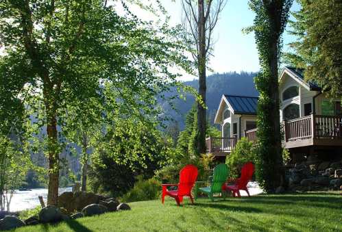 A serene riverside scene with red and green chairs on a grassy lawn, surrounded by trees and mountains in the background.