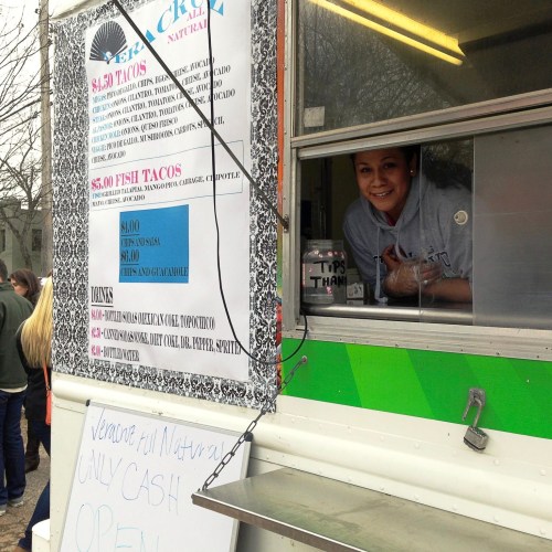 A smiling vendor at a food truck window, offering tacos and drinks with a menu displayed. A sign says "CASH ONLY."