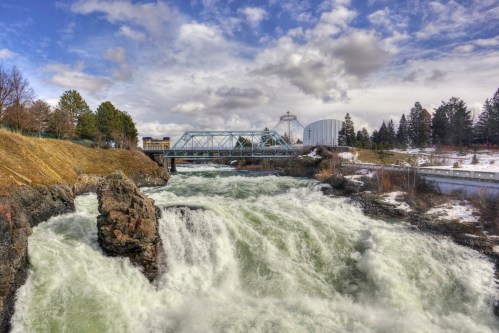 A rushing river flows past a bridge and buildings, surrounded by trees under a cloudy sky.