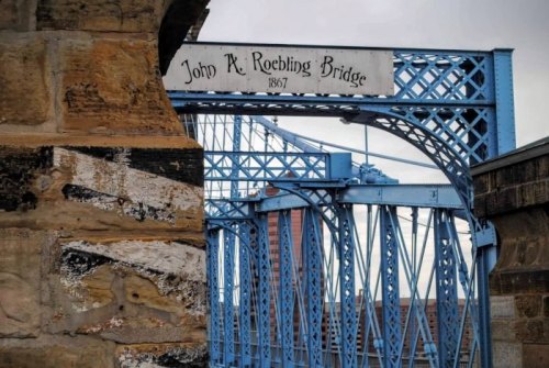Blue steel structure of the John A. Roebling Bridge, featuring a historic sign and stone support.