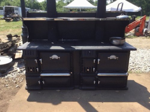 A large, black vintage kitchen stove with multiple burners and ovens, set outdoors on a gravel surface.