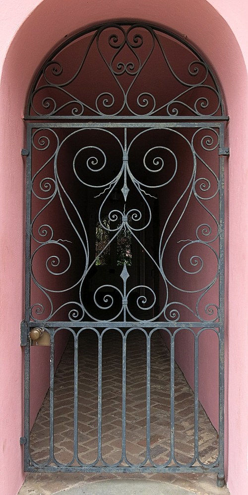 A decorative wrought iron gate with intricate swirls, set against a pink wall, leading into a dimly lit passageway.